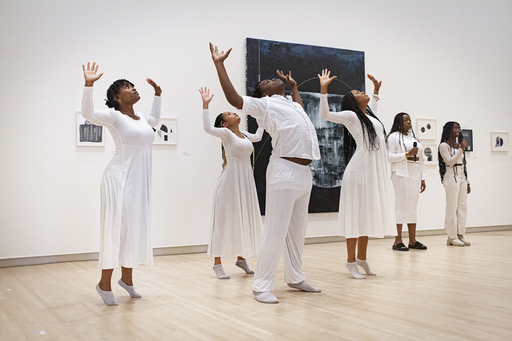 A group of people dressed in white perform in front of artworks hanging on a wall; some are standing with heads back and arms outstretched, while others speak into or hold a microphone