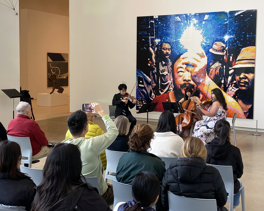 A seated crowd listens as three musicians play stringed instruments in front of Ai Weiwei's artwork "Illumination"