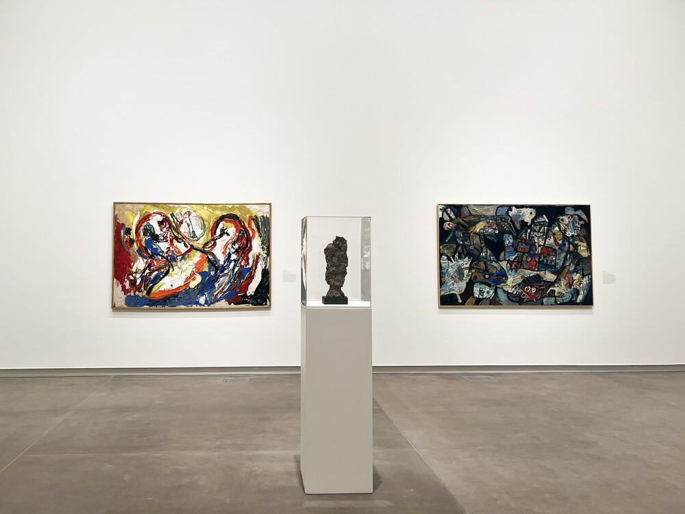 A gallery view with two abstract paintings on a wall, flanking a head-size sculpture in a case