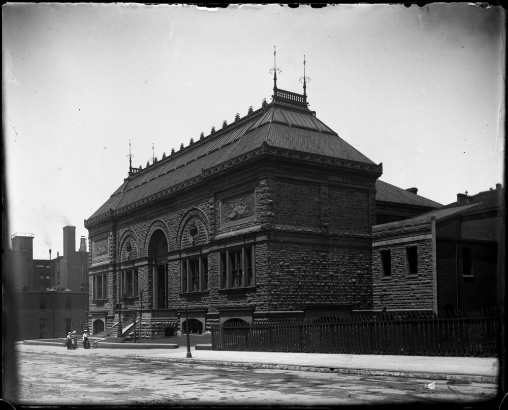 Photograph of the original Museum of Fine Arts building at Nineteenth and Locusts Streets. The picture is taken at a hard angle and shows the side and facade of the building. Three figures in skirts and hats are visible walking in front of the building.