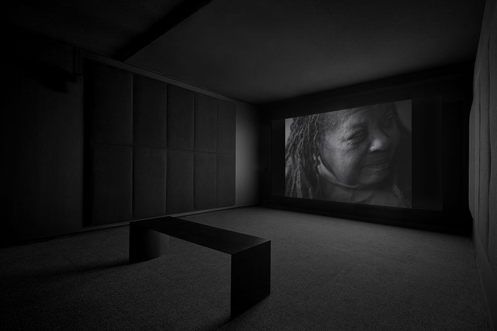 A black-box space with a black bench in the foreground and a black-and-white video on the far wall