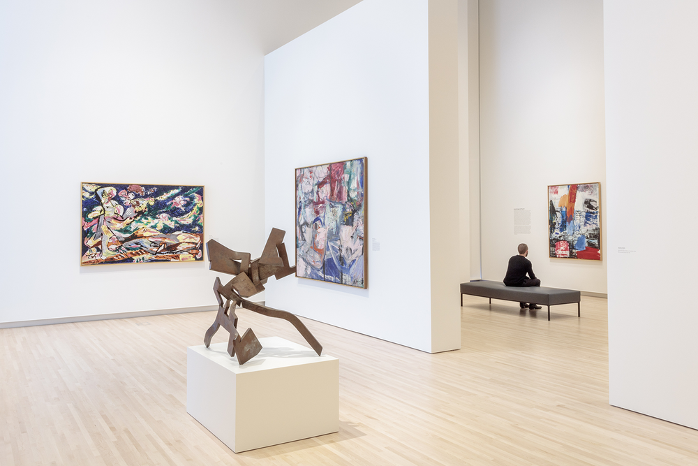 Adjoining galleries displaying abstract paintings on the walls and an abstract sculpture in the foreground; a person is seated on a bench in the far gallery