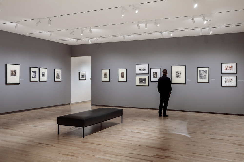 A gallery space with fourteen small framed artworks on the walls; there is a bench in the middle of the room, and a person stands with their back to us looking at an artwork