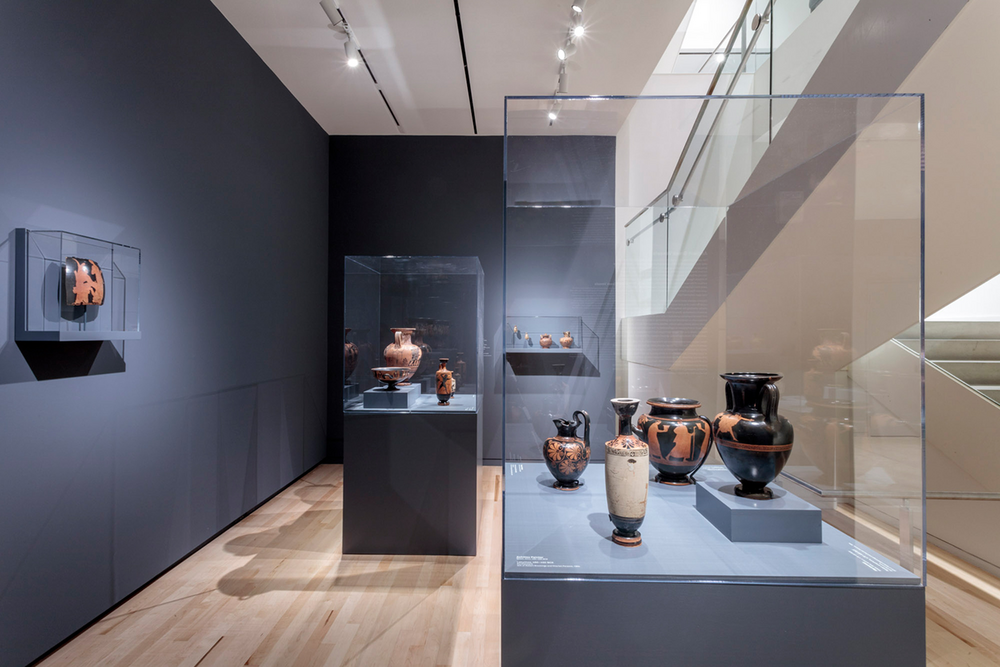 A small gallery space with two large freestanding display cases and two smaller display cases mounted to walls, all containing vases and pottery fragments