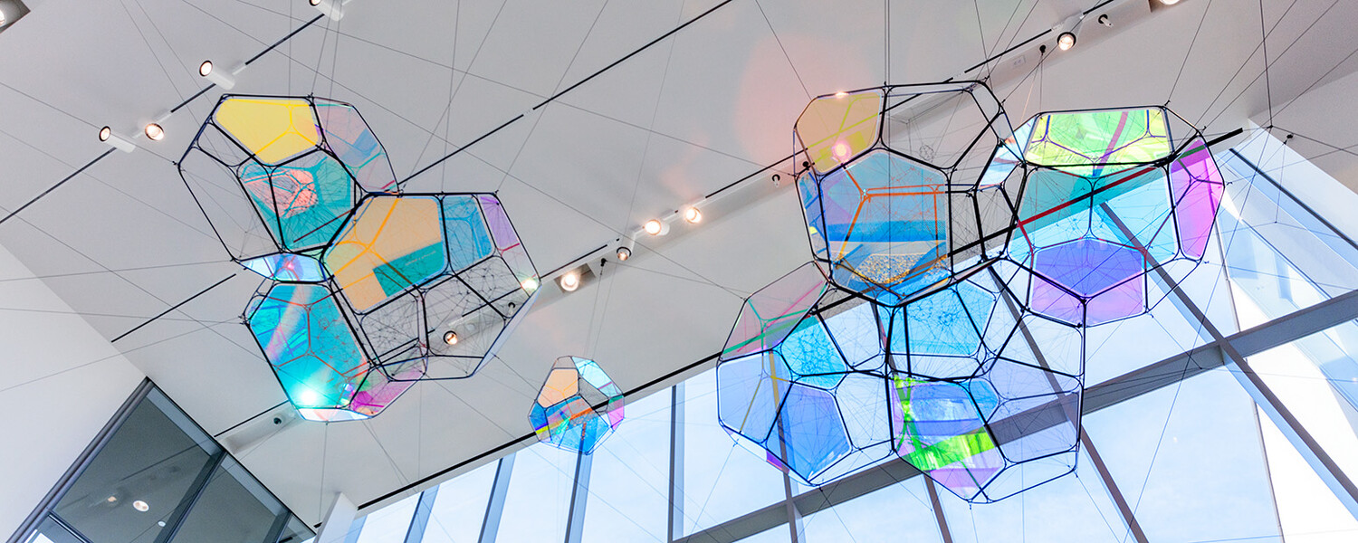 Tomás Saraceno's Cosmic Filaments, a hanging sculptural installation composed of multi-paneled, iridescent modules amid a network of interlocking ropes, viewed from below