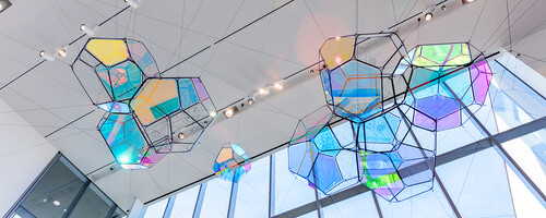 Tomás Saraceno's Cosmic Filaments, a hanging sculptural installation composed of multi-paneled, iridescent modules amid a network of interlocking ropes, viewed from below