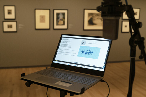 An open laptop sits next to a standing microphone in a gallery with framed prints 