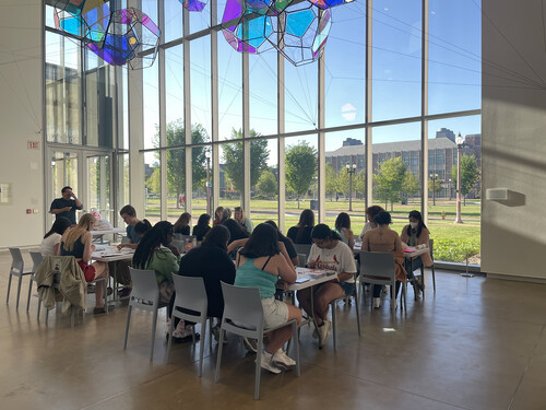 People seated at tables working on a craft project in front of floor-to-ceiling windows and underneath a colorful, iridescent sculpture