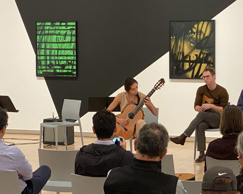 A seated crowd listens as a woman plays guitar in front of two brightly colored paintings