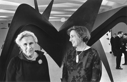 Two women stand in front of a large abstract sculpture