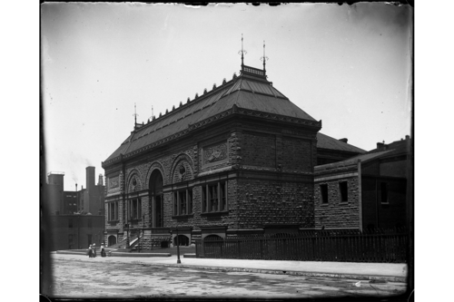 Photograph of the original Museum of Fine Arts building at Nineteenth and Locusts Streets. The picture is taken at a hard angle and shows the side and facade of the building. Three figures in skirts and hats are visible walking in front of the building. 