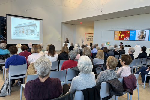 A group of people seated in the museum's lobby looking at an image of the old museum building on a projector screen.