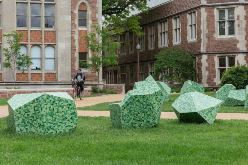 A sculpture outdoors on grass. There is a person biking on the path behind the sculpture.